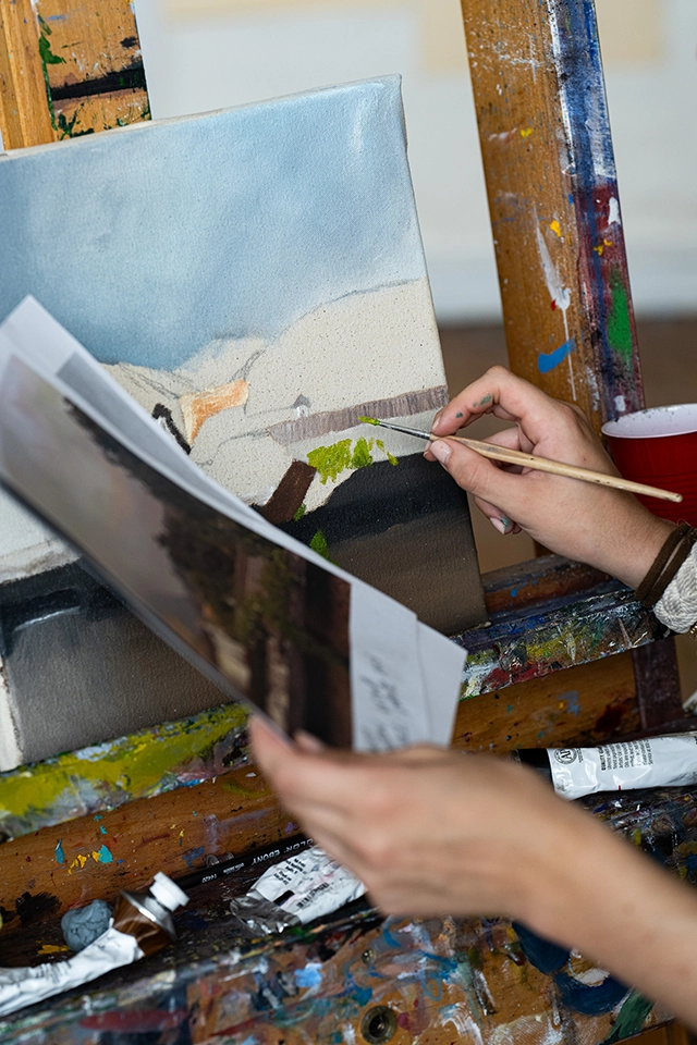 A person paints a landscape on canvas using a brush, referencing a photo held in their other hand. Various art supplies and paint tubes are on the easel.