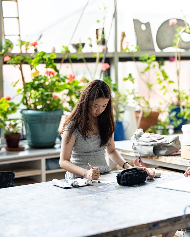 A woman with long hair works on an art project at a table in a bright room filled with plants and art supplies. Sunlight streams through the large windows behind her.