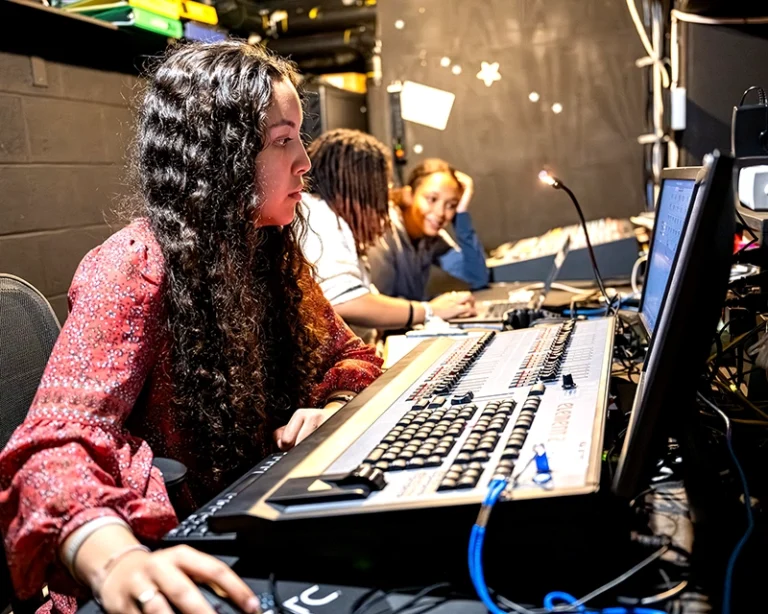 A woman with long curly hair operates a control panel backstage, focusing on a monitor, as two other people work beside her in a dimly lit room filled with electronic equipment.