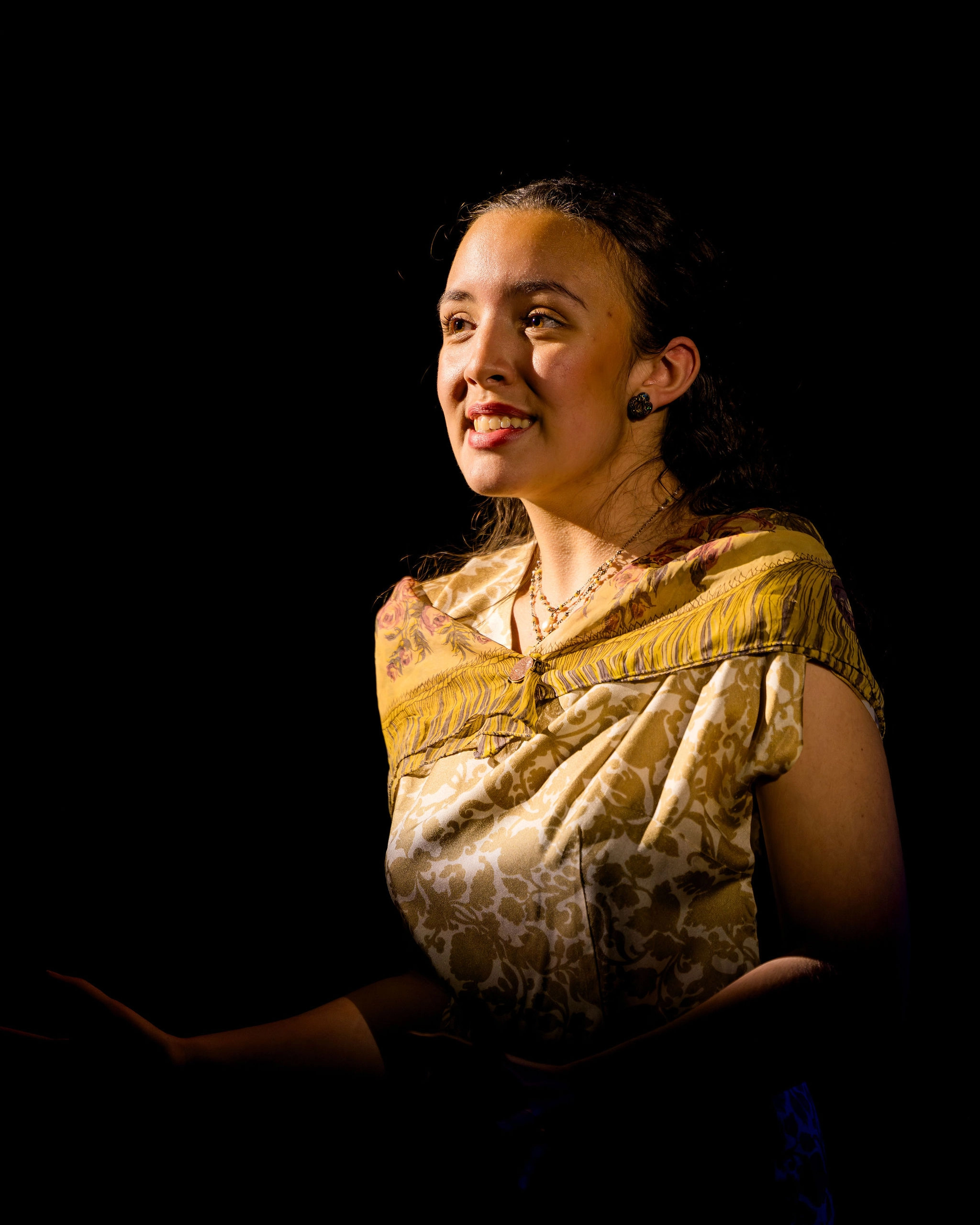 A young woman wearing a patterned dress and shawl is smiling, lit by a spotlight against a black background.