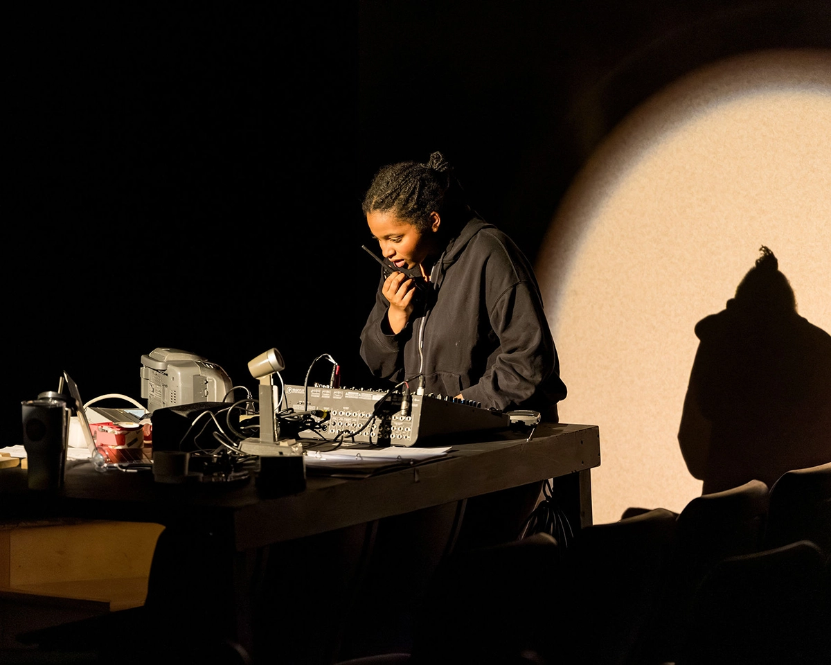 A person in a dark hoodie stands at a table with audio equipment, holding a walkie-talkie to their mouth. A spotlight creates their shadow on the wall behind them.