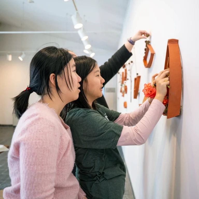 Two women closely examine and adjust orange decorations with flowers on a white gallery wall under bright ceiling lights.