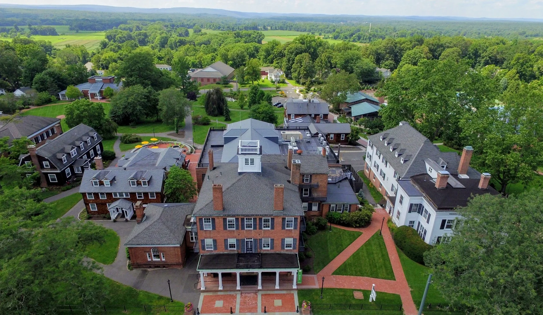 Aerial view of Miss Porter's School campus with historic brick and white buildings, green lawns, pathways, and surrounding trees, set against a backdrop of fields and distant hills under a partly cloudy sky.