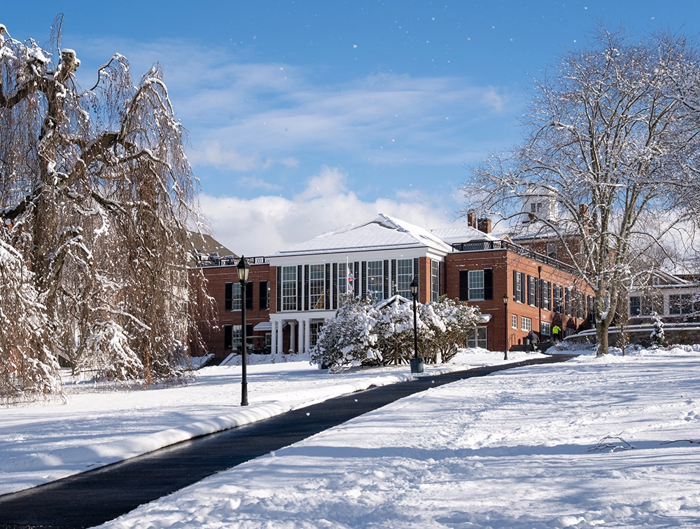 A red-brick building with large windows stands at the end of a snow-covered path, surrounded by snowy trees under a bright blue sky.