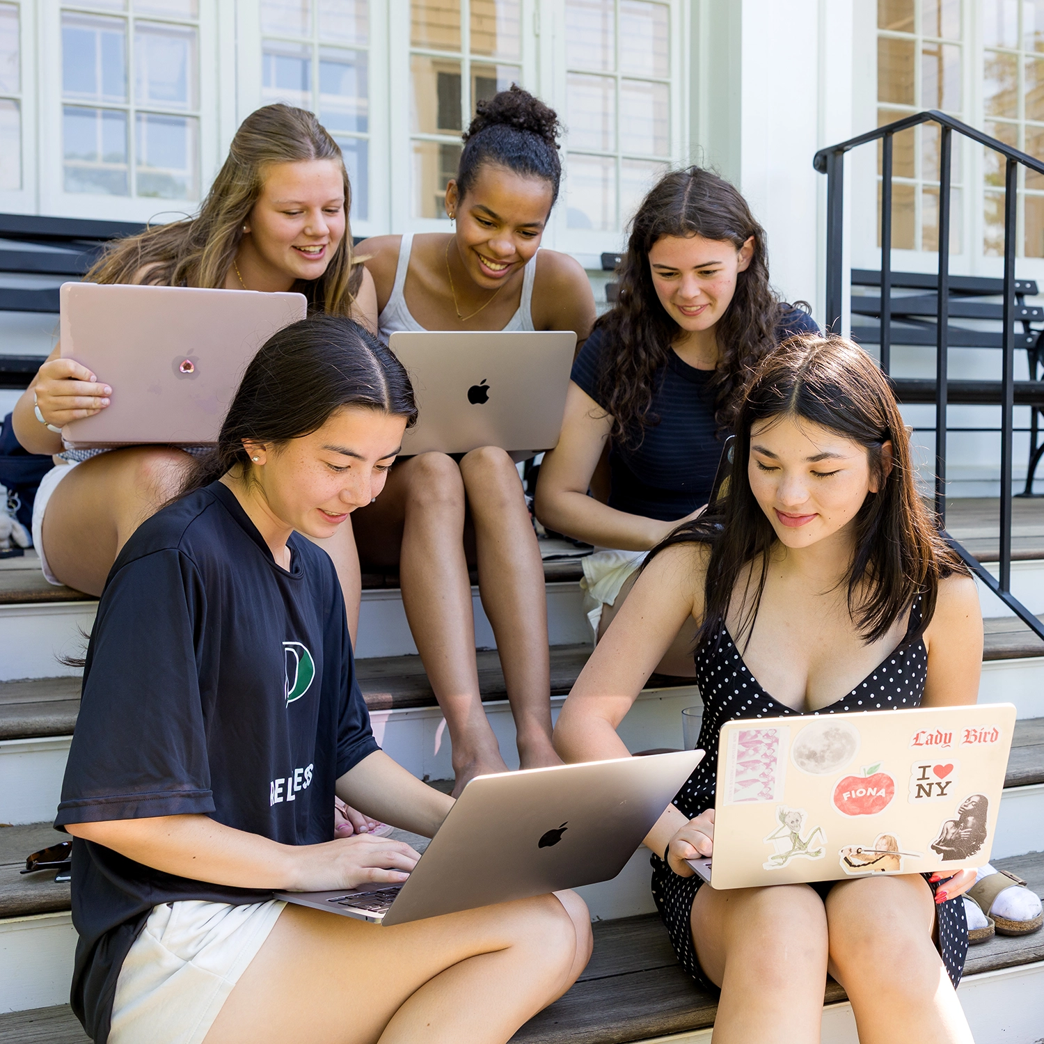 Five young women sit on outdoor steps, smiling and working on laptops together. The background shows white railings and windows, suggesting a school or campus setting.