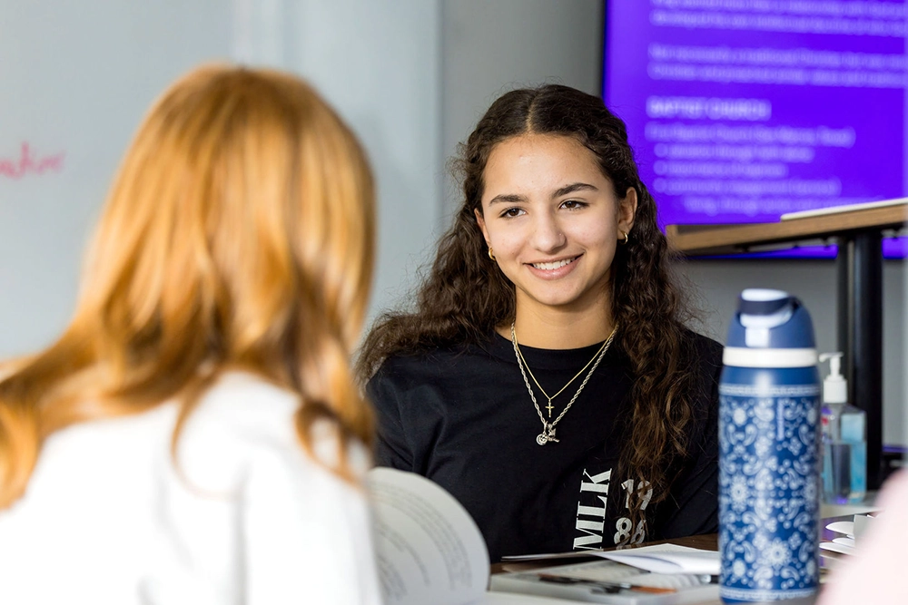 Two students sit at a table, talking and smiling. One girl with long curly hair faces the camera, while the other, with reddish hair, has her back to the camera. A blue water bottle sits on the table.