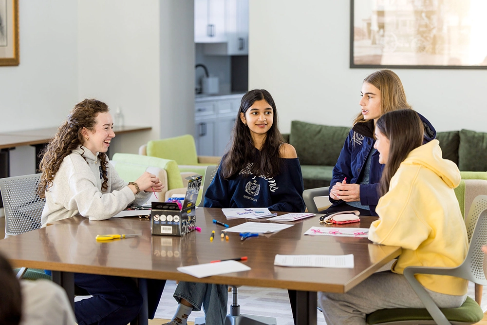 Four teenage girls sit around a table in a bright room, talking and smiling while working on school assignments with papers, pencils, and a laptop.
