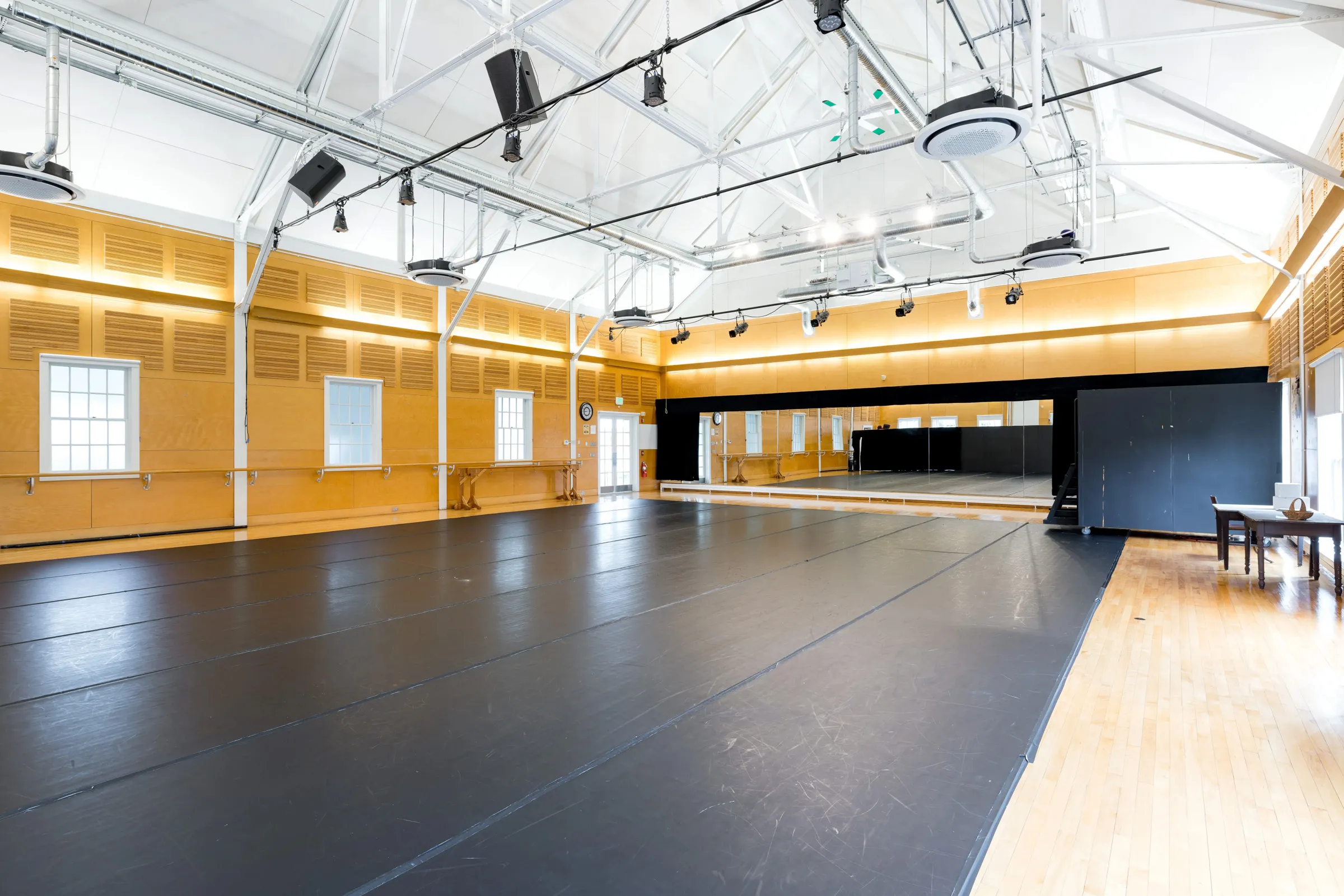 A spacious, bright dance studio with a black marley floor, large mirrors along one wall, high ceilings, and tall windows letting in natural light. There is a barre, a clock, and a table with two chairs in the corner.