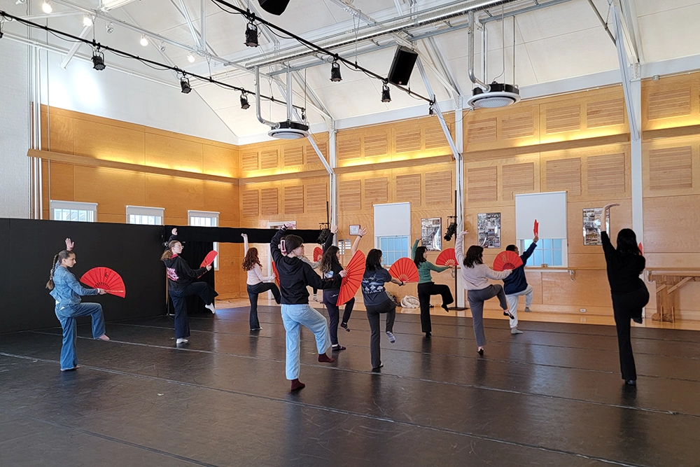 A group of people in a dance studio practice with red fans, each raising one leg and holding the fan up. The room has wooden walls, large windows, and overhead lights.