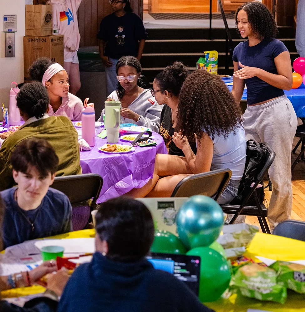 A group of people sit and stand around tables covered with purple cloths, eating and talking. The tables have food, drinks, balloons, and art supplies. The atmosphere appears lively and social.