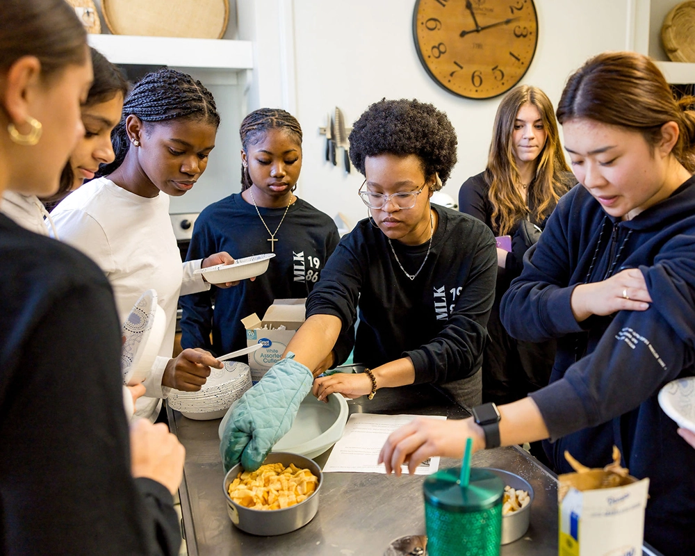 A group of young people gather around a kitchen counter, preparing food together. One person wears an oven mitt and handles a dish, while others watch or help. A large clock hangs on the wall in the background.