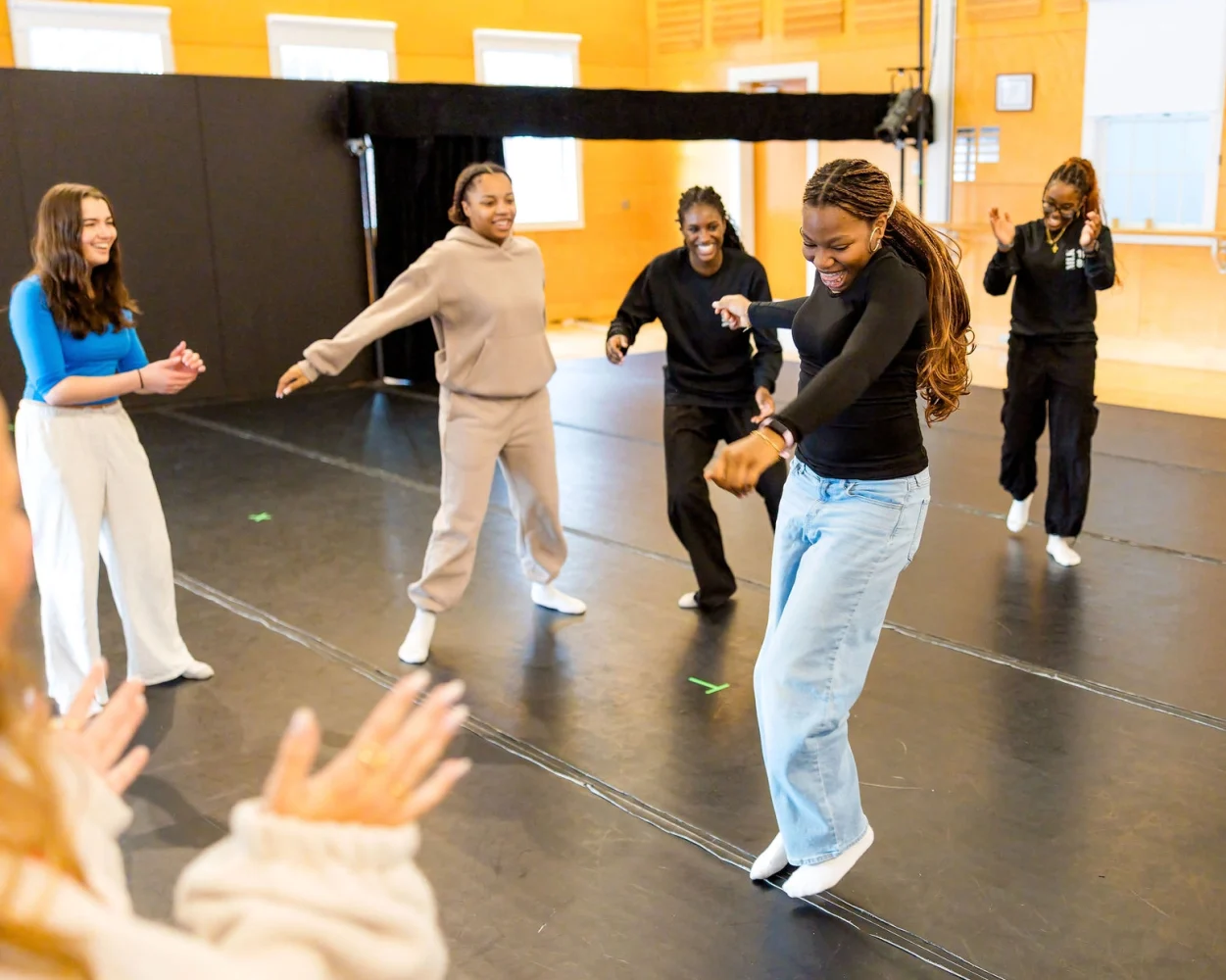 Five people smile and dance together in a brightly lit room with wooden walls, while two others applaud in the foreground. Everyone appears energetic and joyful as they move and interact.