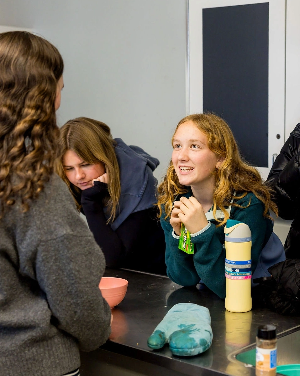 Three girls sit at a desk near a bowl and bottles. One girl smiles and talks to another, while a third girl behind them leans on the table, looking tired or distracted.