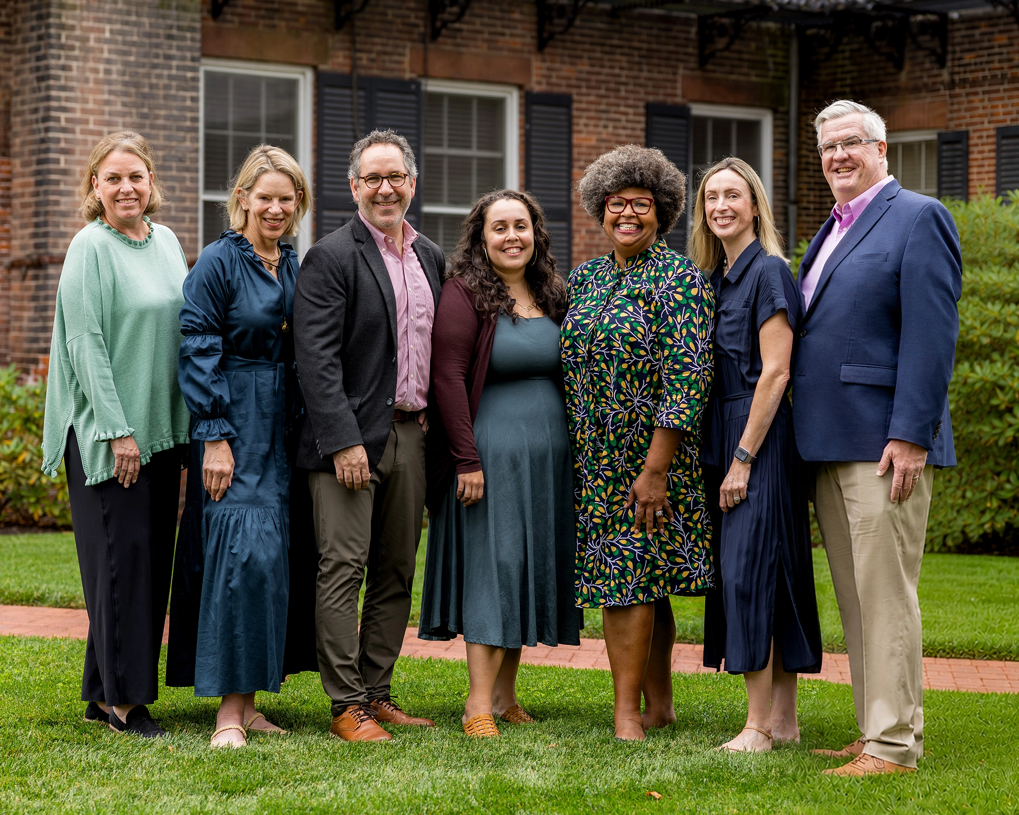 The MPS leadership team stand side by side outdoors on grass, smiling at the camera. They are dressed in semi-formal attire, with greenery and a brick building in the background.