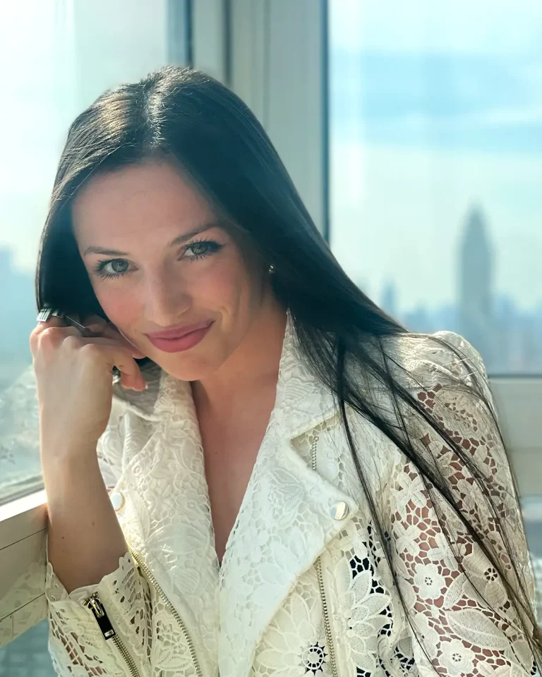 A woman with long dark hair and light eyes smiles softly while leaning on her hand by a window, wearing a white lace jacket. The city skyline is visible in the blurred background.