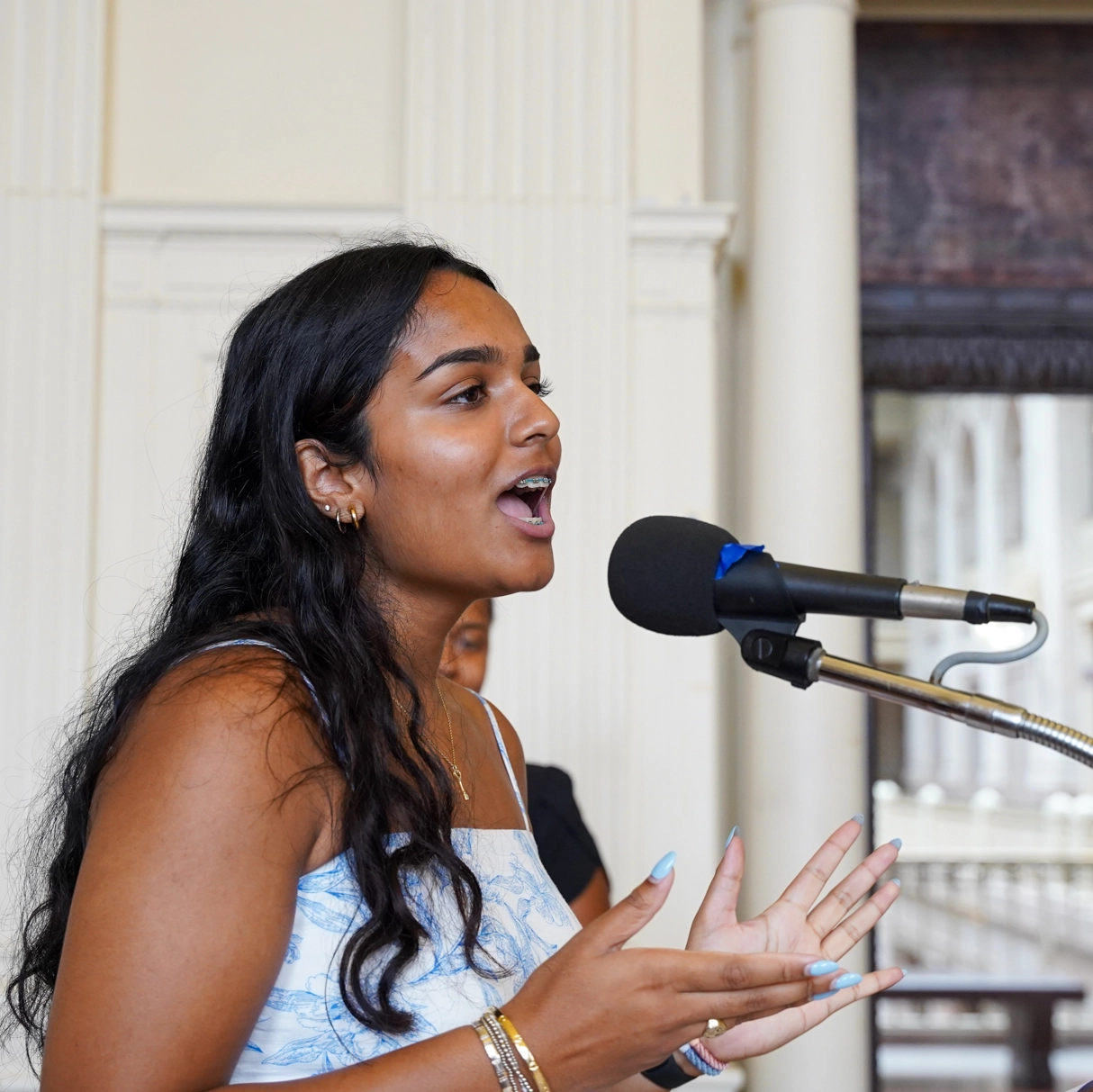 A woman with long dark hair speaks passionately into a microphone, gesturing with her hands. She wears a white and blue dress and gold jewelry, standing indoors with columns and large windows in the background.