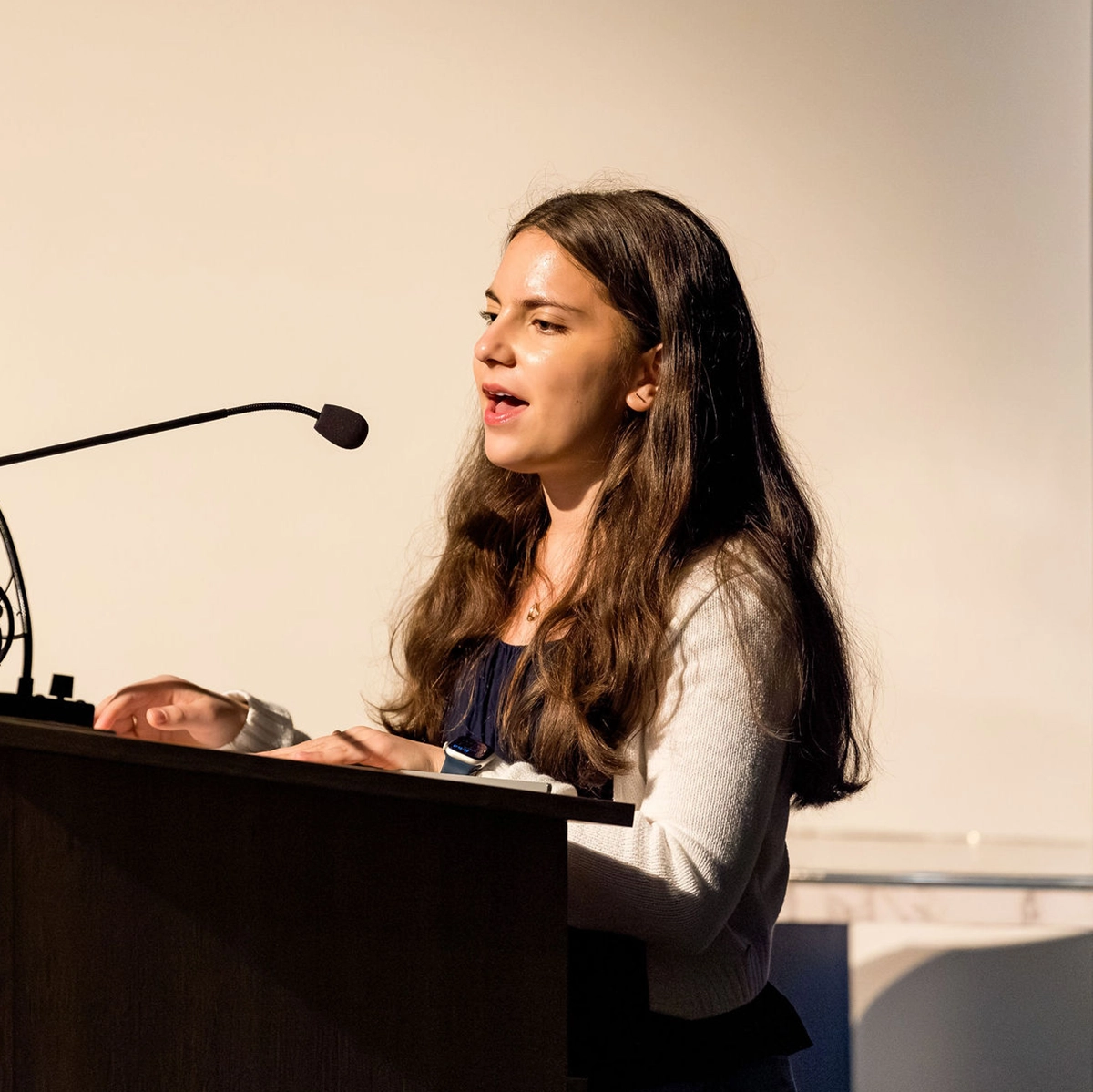A young woman with long brown hair speaks into a microphone while standing at a podium. She appears to be giving a speech or presentation in a well-lit indoor setting.