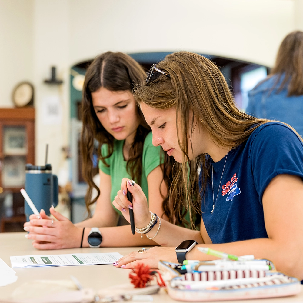 Two young women sit at a table, focused on a worksheet. One writes while the other observes. Pens, paper, and a drink are on the table. The background is a blurred indoor setting.