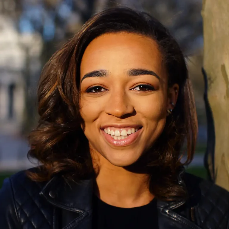 A woman with medium-length brown hair smiles at the camera while standing outdoors. She wears a black quilted leather jacket and a black top. Sunlight illuminates her face, and trees are visible in the blurry background.