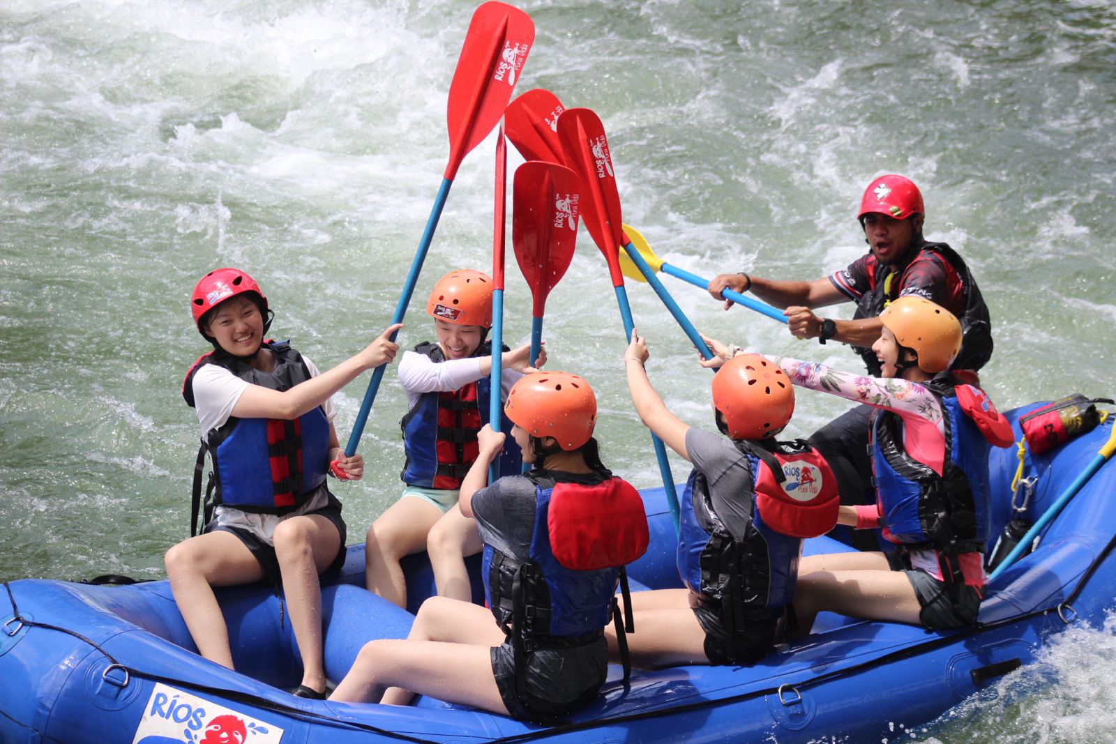 A group of six people wearing helmets and life jackets high-five with paddles while whitewater rafting in a blue inflatable raft on a river. The guide steers from the back.
