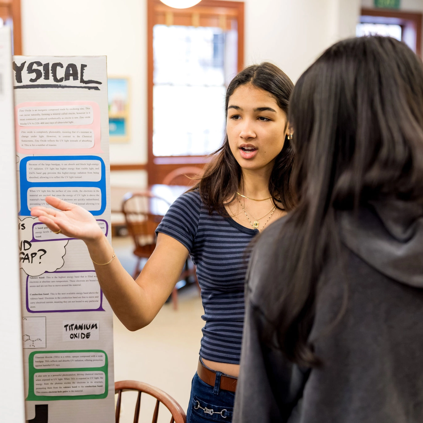 A young woman presents a science project display board to another person in a classroom. She gestures with her hand while explaining, and the display board contains colorful text boxes and diagrams.