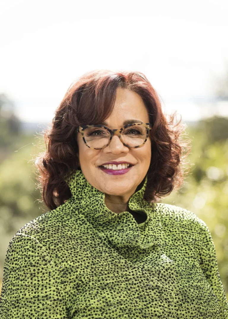 A woman with shoulder-length curly hair, wearing glasses and a green patterned high-neck top, smiles outdoors with a blurred, sunny background of greenery.