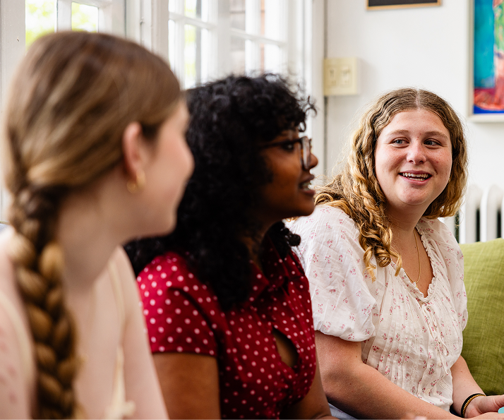 Three young women sit together on a couch, talking and smiling. The woman on the right looks at the others with a friendly expression. Bright natural light enters through windows behind them.