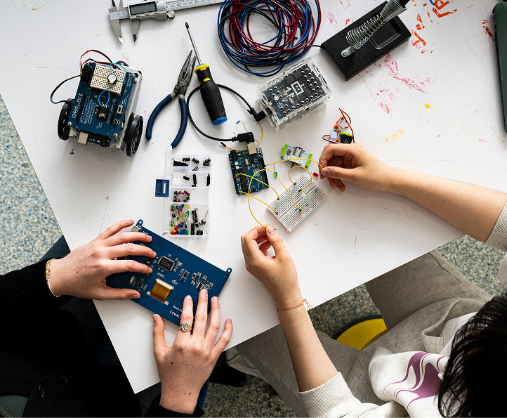 Two people work on electronics projects at a table, using circuit boards, wires, and tools such as pliers and a screwdriver. Various electronic components and a small robot are also visible on the table.