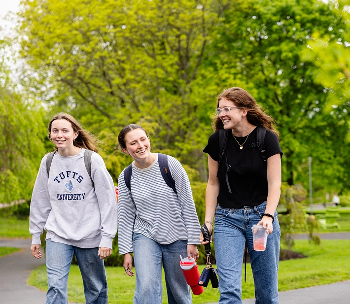 Three young women wearing casual clothes and backpacks walk together outside on a green, tree-lined path, smiling and talking. One holds a drink, another carries a red bottle. It looks like spring or summer.