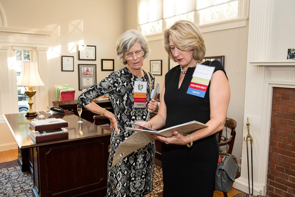 Two women stand in an office, examining a document together. Both wear name tags and conference ribbons. Sunlight streams through windows, and office furnishings are visible in the background.