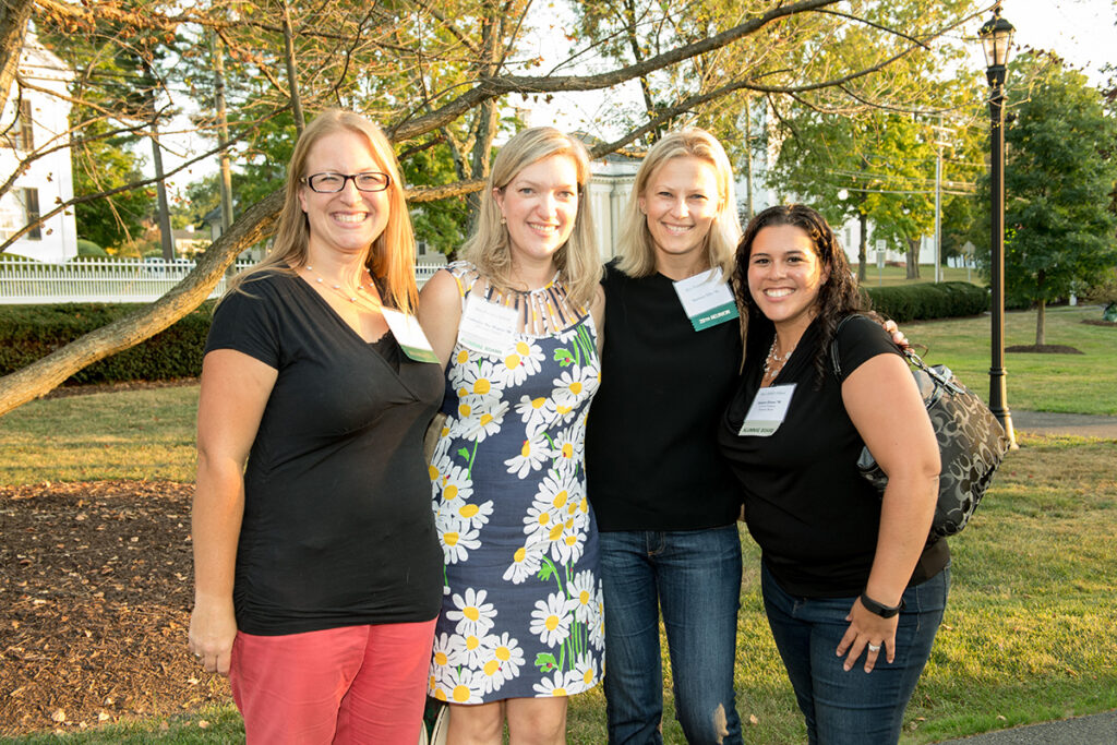 Four women stand close together outside, smiling at the camera. Three wear name tags. Trees, grass, and a lamp post are in the background, suggesting a park or campus setting. It appears to be sunny.