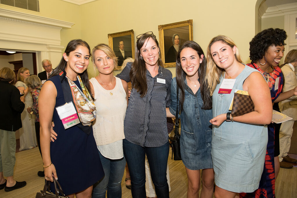 Five women posing together and smiling at an indoor social event, with other people and portraits on the wall in the background.