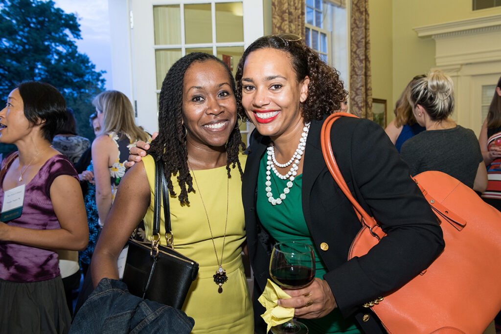 Two women smile at a social event, one in a yellow dress and the other with a black blazer and orange bag, holding a glass of wine. People chat in the background in a brightly lit room.