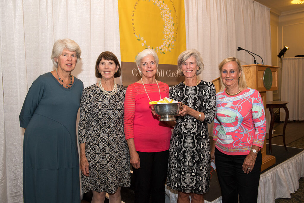 Five women stand together in front of a yellow banner. One woman holds a metal bowl filled with yellow papers. They are dressed in semi-formal attire and are posing for a group photo at an indoor event.