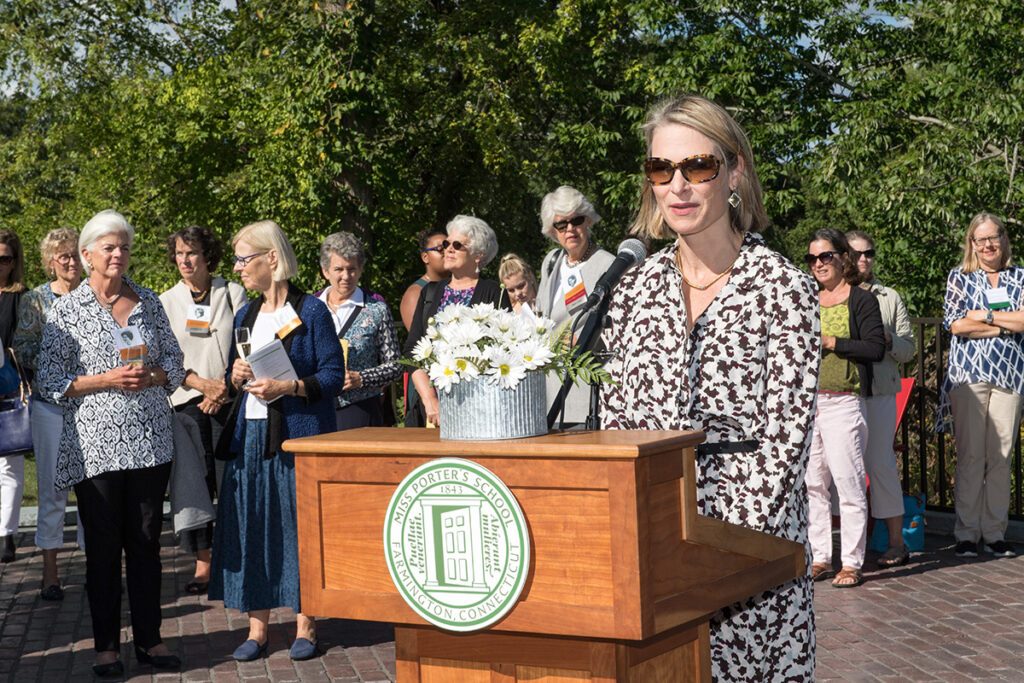 A woman speaks at a podium outdoors, wearing sunglasses and a patterned dress. A group of people stand behind her, listening. The podium has a bouquet of flowers and a green emblem on the front. Trees are in the background.
