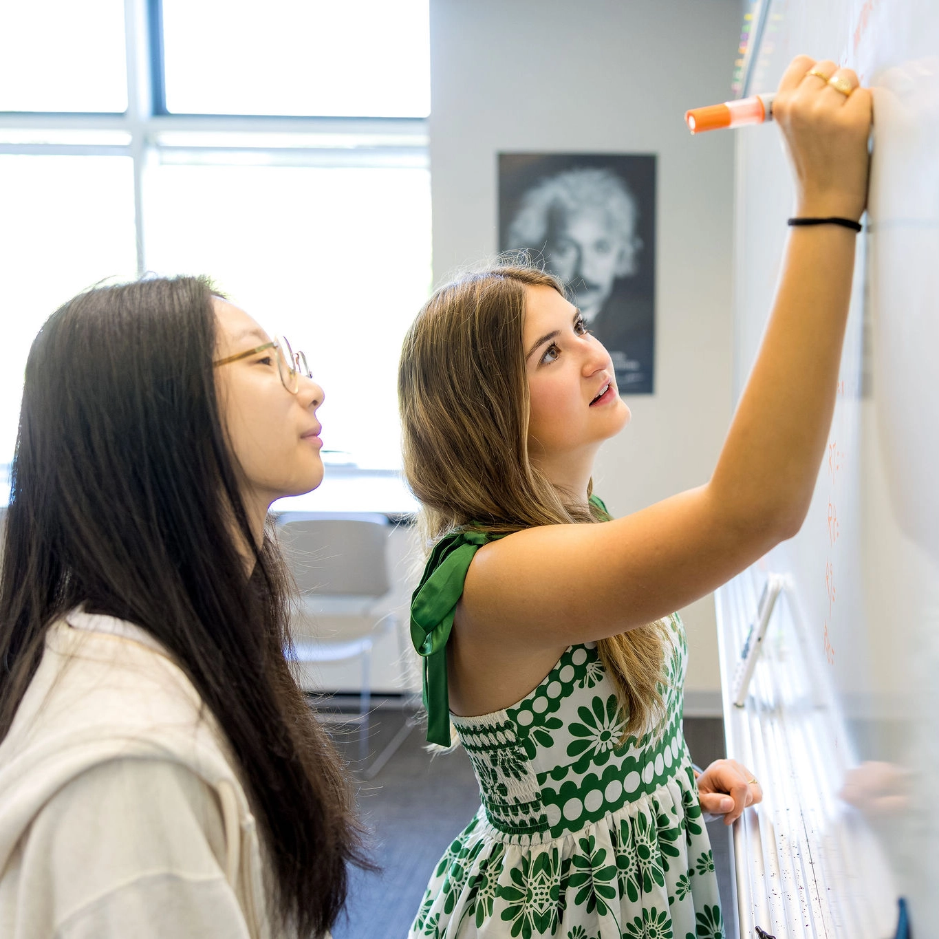 Two young women work together at a whiteboard, one writing with an orange marker while the other observes. A black-and-white portrait is blurred in the background. Sunlight streams through large windows behind them.
