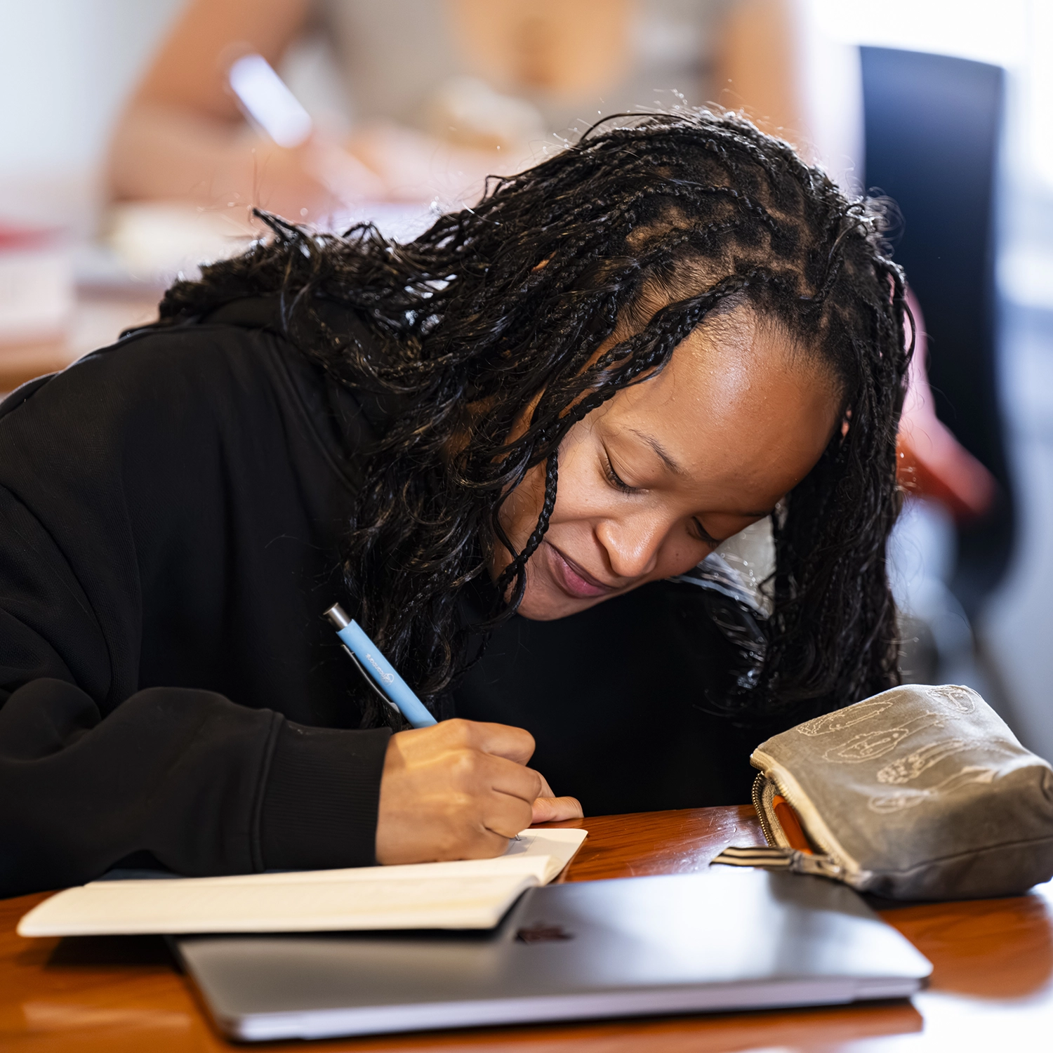 A person with braided hair writes in a notebook with a blue pen while sitting at a desk. A pencil case and a closed laptop are nearby. Another person is blurred in the background.