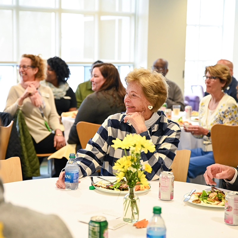 A group of people sit around tables eating and talking at a daytime indoor event. A smiling older woman in a striped shirt is in focus, holding a water bottle. Yellow flowers decorate the tables.