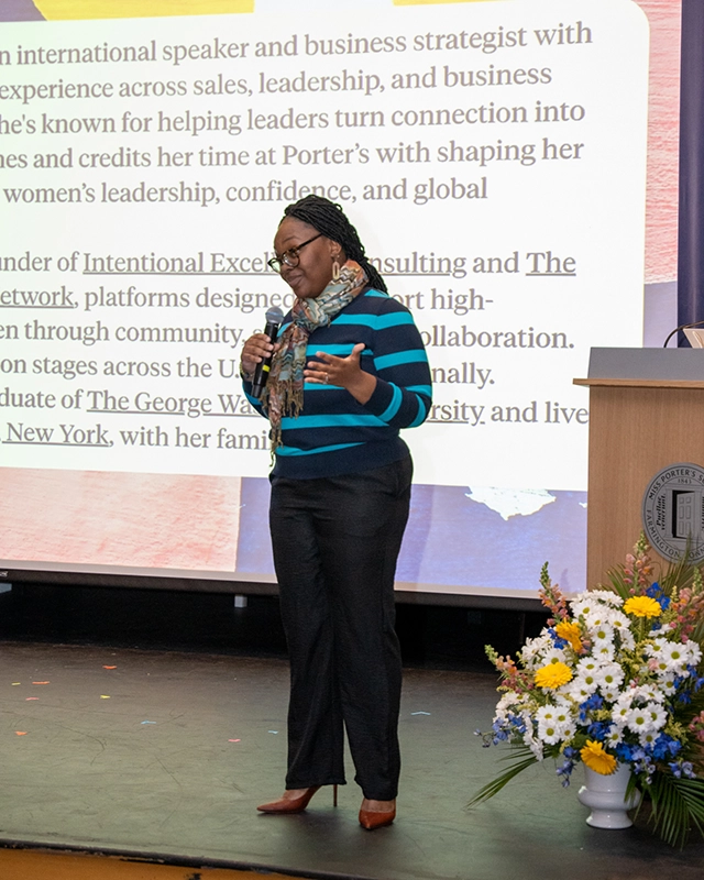 A woman wearing glasses and a striped sweater speaks on stage with a microphone. Behind her, a large screen displays text. A podium and a colorful flower arrangement are also visible.