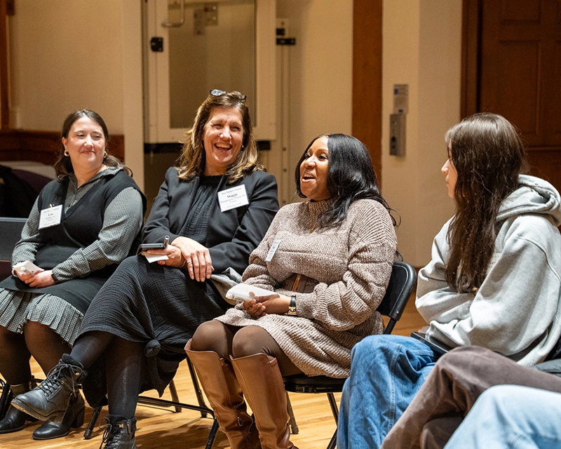 Four women sit in chairs indoors, smiling and laughing together during a group discussion. They wear name tags and casual to business-casual clothing. The setting appears warm and collegial.