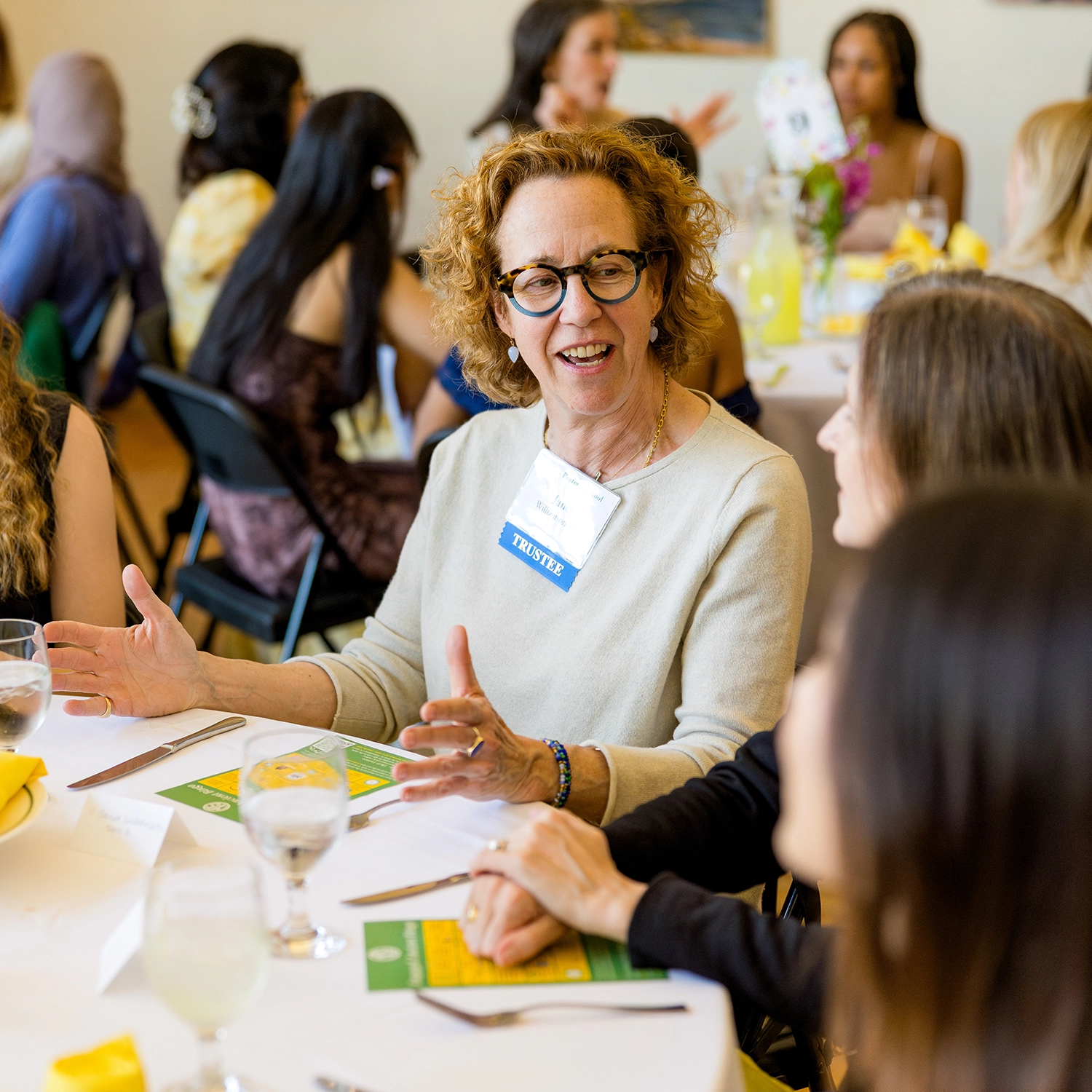 A woman wearing glasses and a Trustee name tag gestures while talking to others at a round table set with drinks, menus, and yellow napkins during a social event.