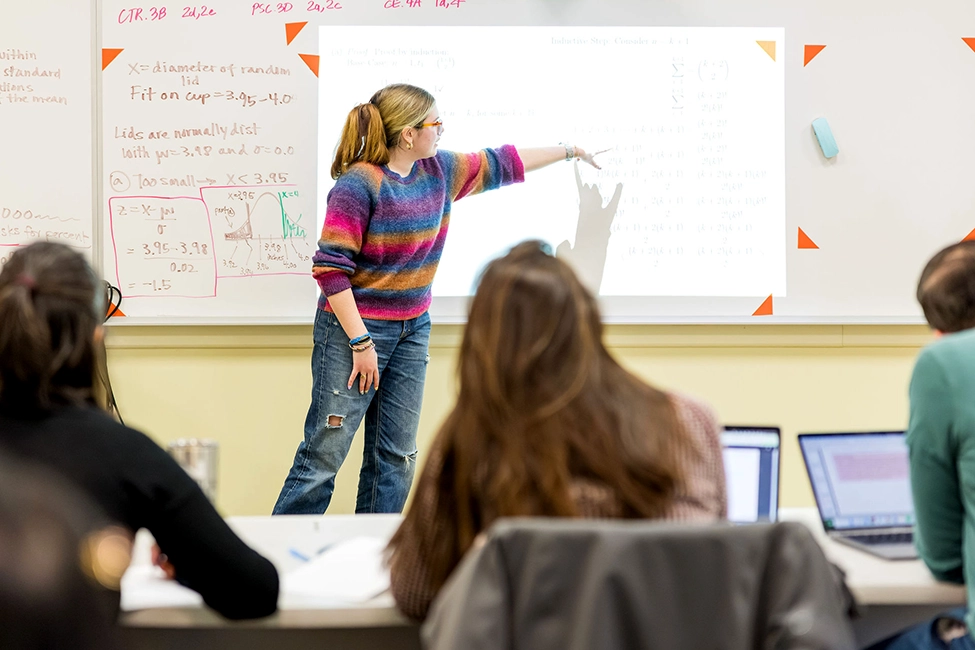 A person in a colorful sweater stands at the front of a classroom, pointing at a projected screen, while students sit at desks with laptops and notebooks, facing the presenter.