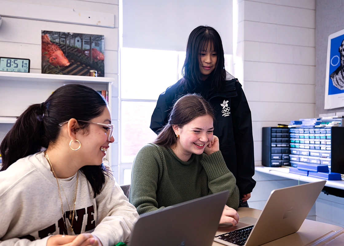 Three young women in a classroom work together on laptops, smiling and talking. One stands, while the other two sit at a table. Books, shelves, and a clock are visible in the background.