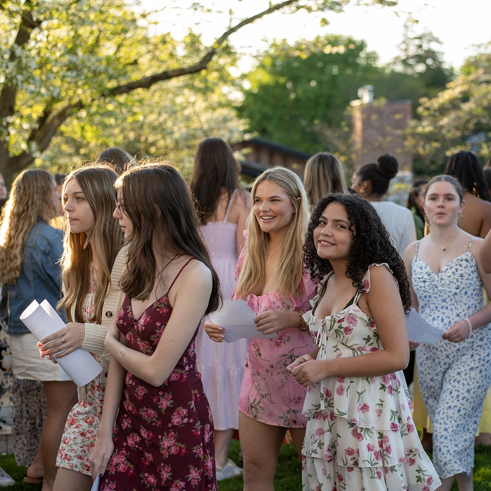 A group of young women in floral dresses stand outdoors, smiling and holding papers, with sunlight filtering through the trees in the background.