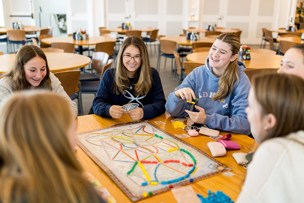 Five young women sit around a table in a bright room, smiling and playing a board game with colored pieces and cards. Empty tables and chairs are visible in the background.