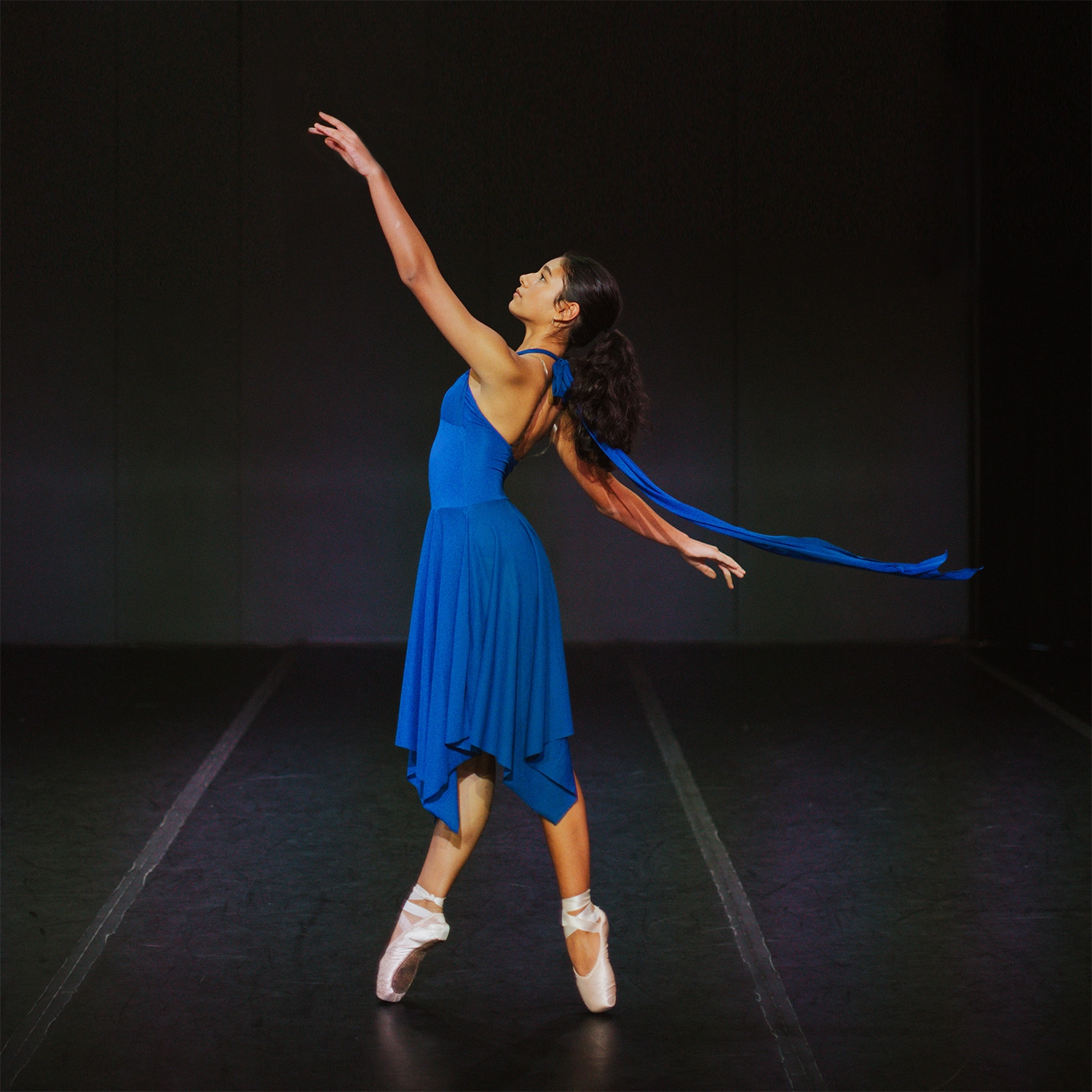 A ballet dancer in a flowing blue dress performs en pointe on stage, with one arm reaching upward and a blue ribbon trailing behind her.