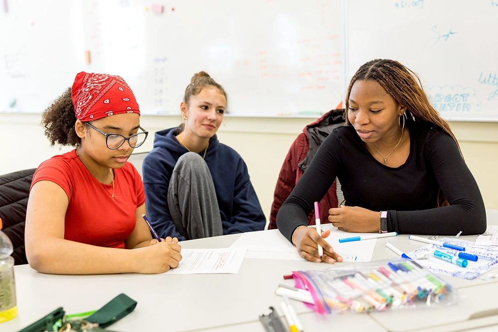 Three students sit at a table in a classroom, writing on paper. One wears a red bandana, another sits with knees up, and the third wears a black shirt. Pens and markers are scattered on the table.