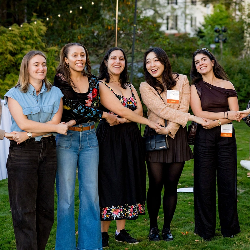 Five women stand outdoors on grass, linking arms and smiling. They wear casual and semi-formal outfits, and several have name tags. Trees and string lights are visible in the background.