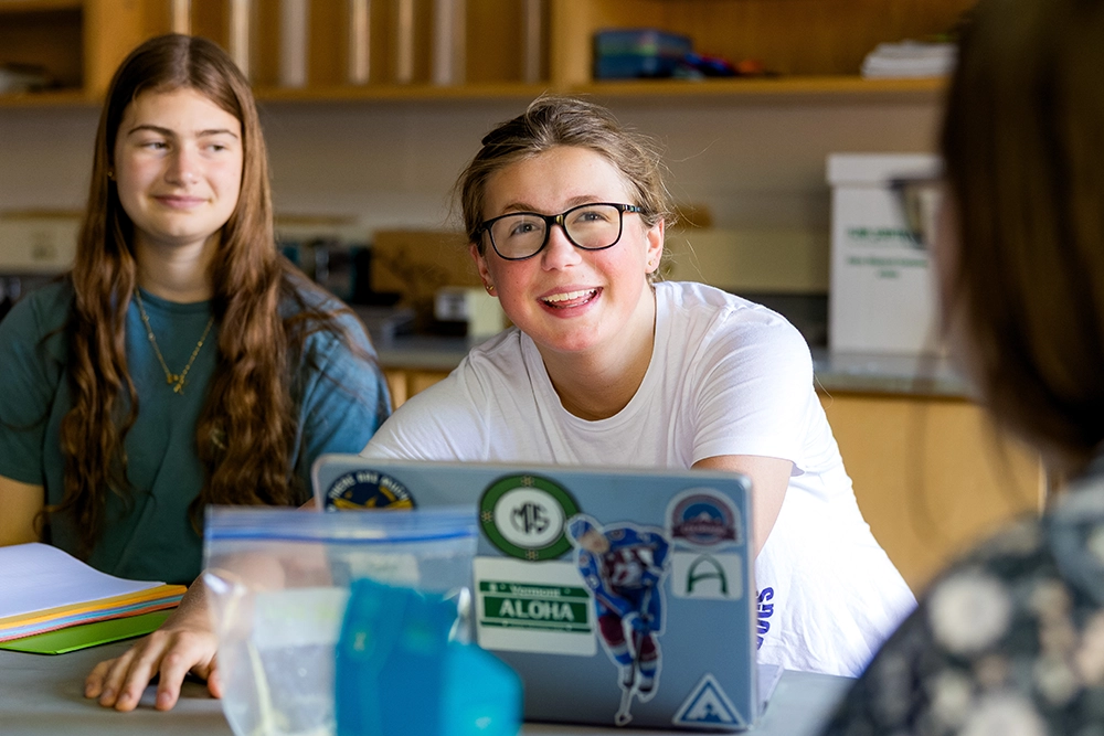 Two young women sit at a table in a classroom. One, wearing glasses and a white shirt, smiles while facing someone off-camera. A laptop covered in stickers sits in front of her; papers and folders are on the table.