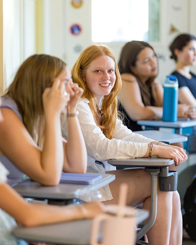 A group of students sit at desks in a classroom. One student with long red hair, wearing a white shirt, sits in focus and smiles at the camera. Others are blurred in the background.